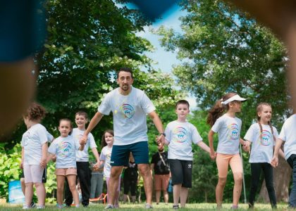 Children and an instructor playing together in a sunny park, holding hands and enjoying outdoor activities.